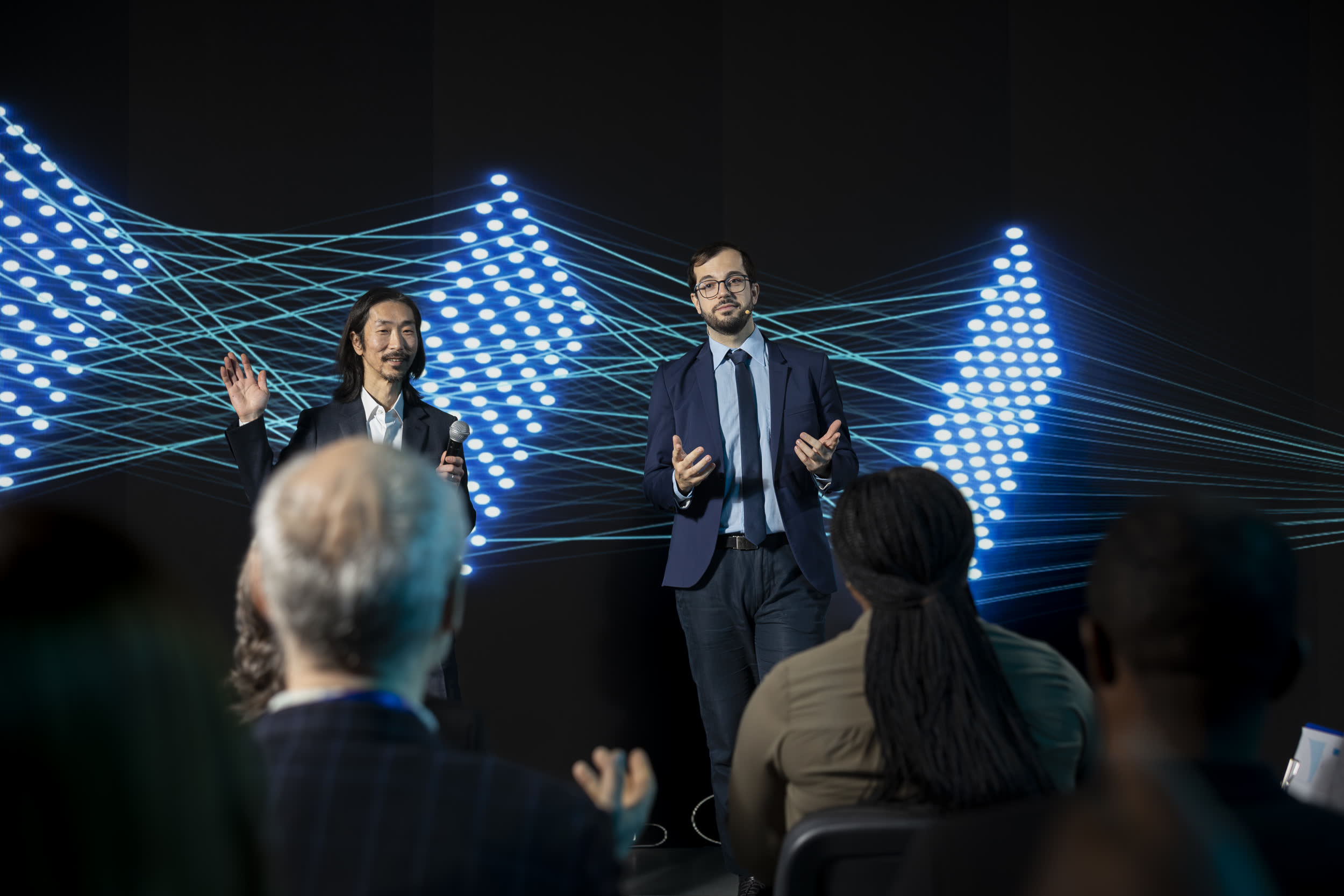 Two speakers presenting on a conference stage lit by LED panels